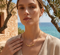 Woman wearing a gold necklace with a scenic background of ocean and stone building.