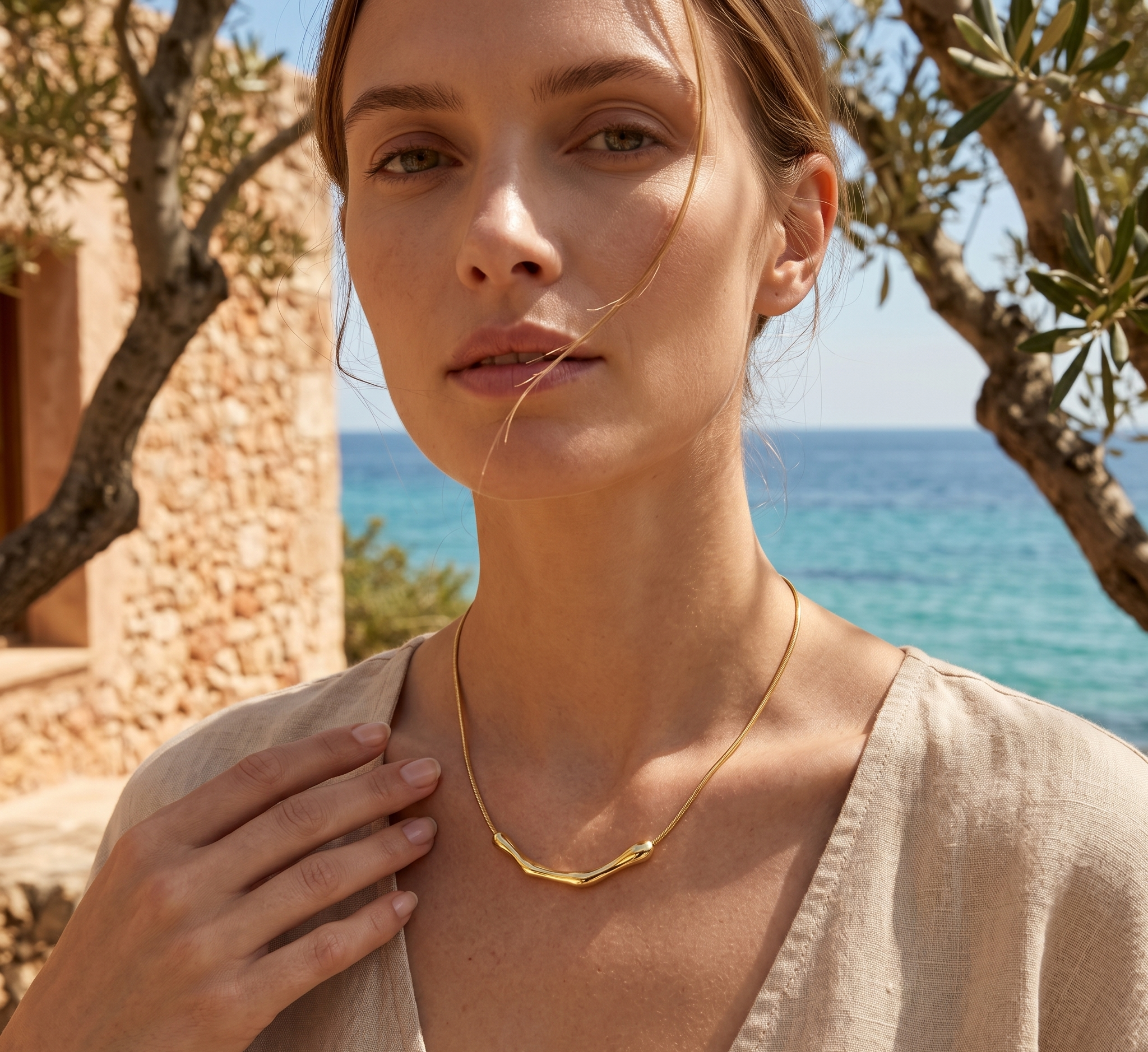 Woman wearing a gold necklace with a scenic background of ocean and stone building.